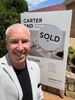 Smiling man stands beside a real estate sign with a large “SOLD” notice.