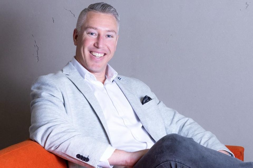 Smiling man in a light gray blazer seated in an orange chair against a plain wall