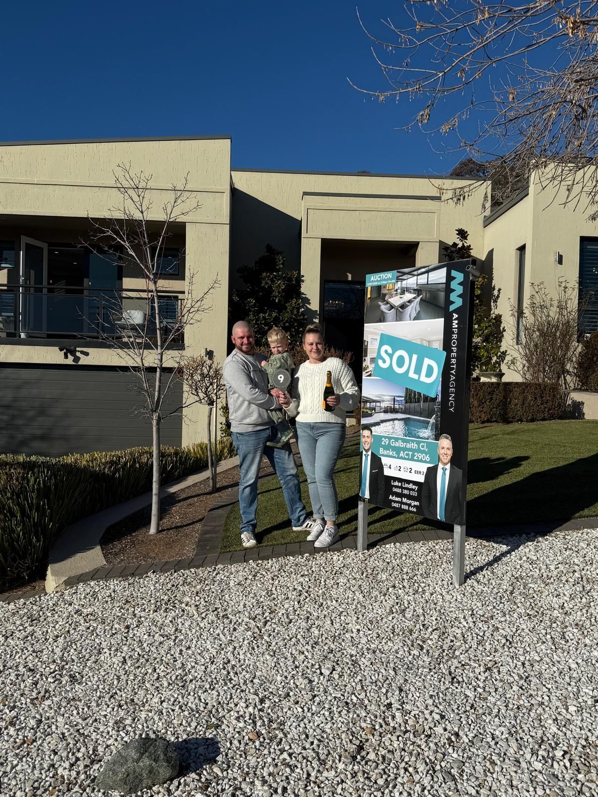 Three people posing beside a real estate “SOLD” sign outside a modern building.