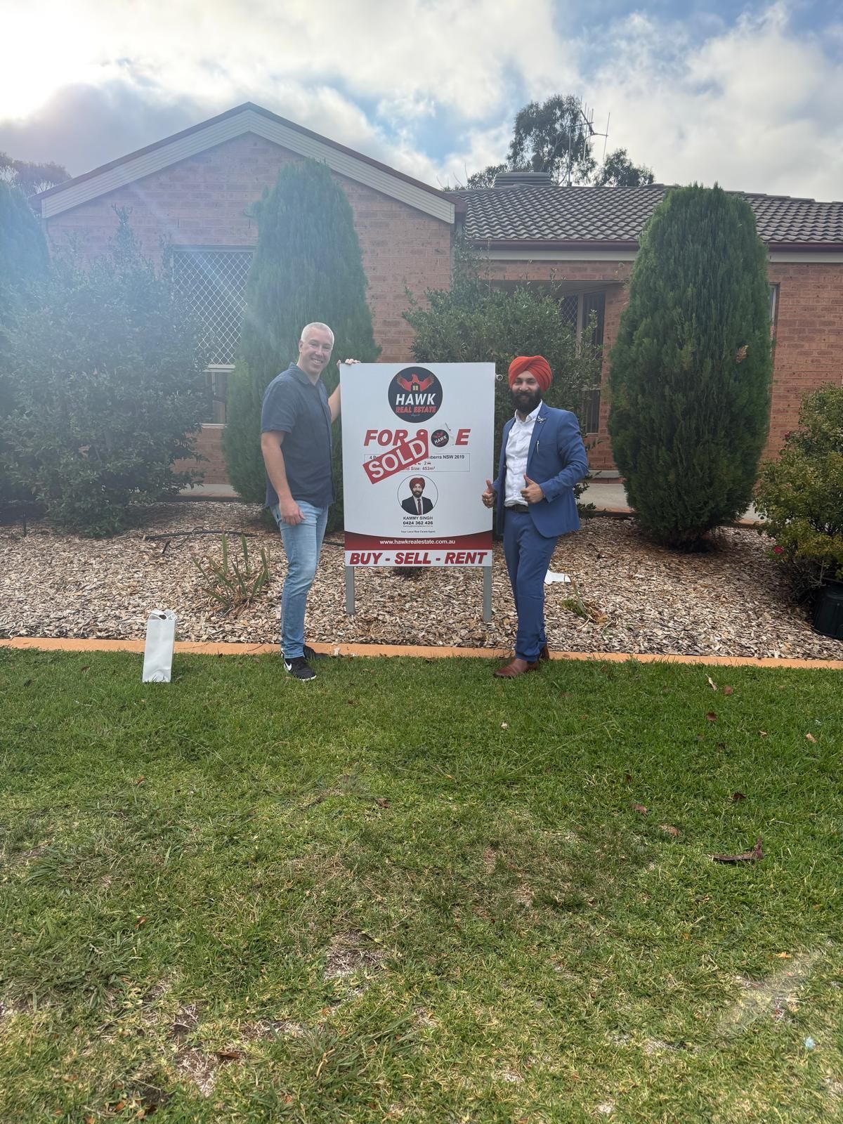 Two people pose beside a sign in front of a house and rock garden.