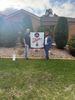 Two people pose beside a sign in front of a house and rock garden.