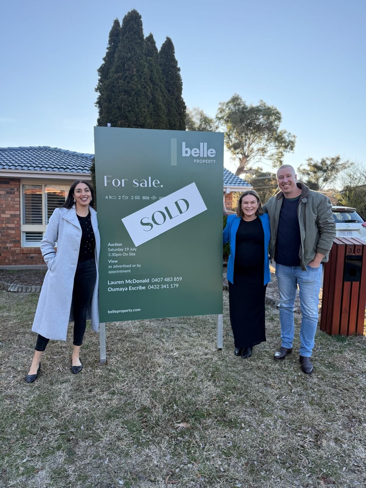 Three people standing beside a house-for-sale sign marked “SOLD” in front of a home.