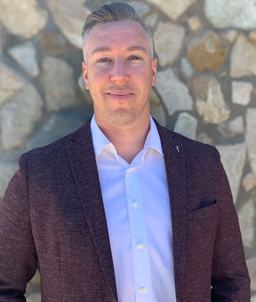 Man in a maroon blazer and white shirt standing against a stone wall, smiling outdoors