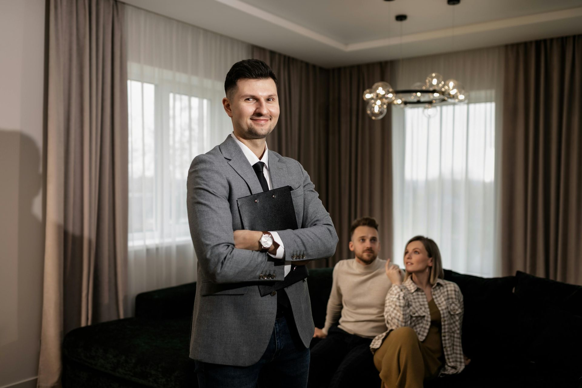 Man standing with arms crossed in a living room, with two people seated on a sofa behind him.