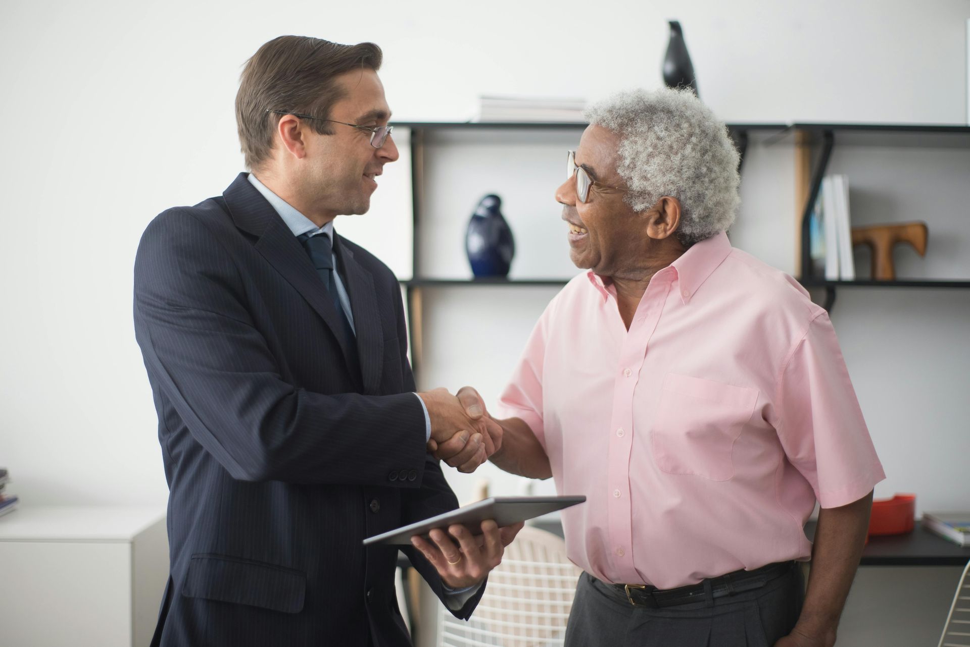Two professionals shaking hands in an office, one holding a tablet.