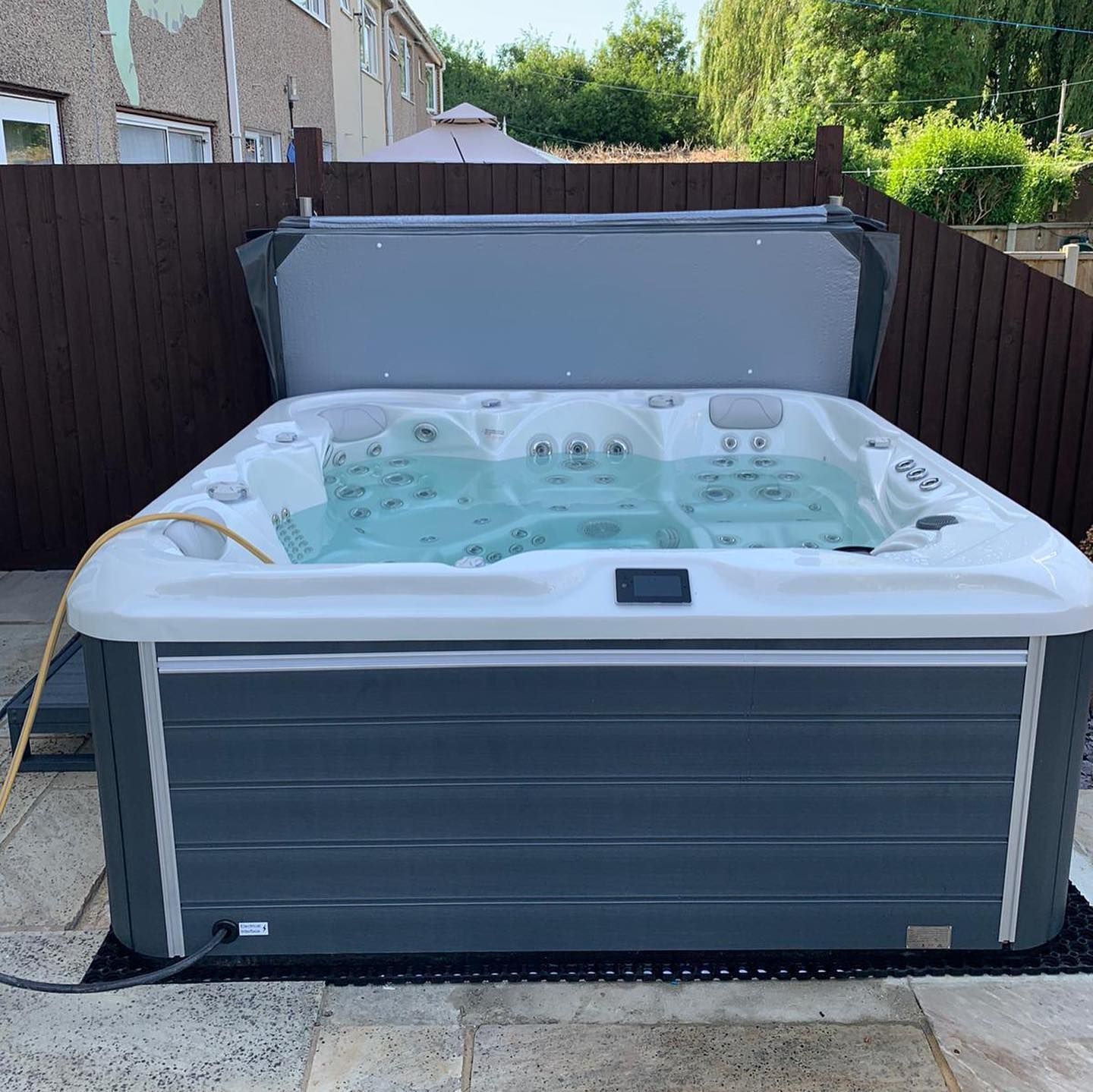 Hot tub, gray and white, with open cover, set on patio, surrounded by a wooden fence.