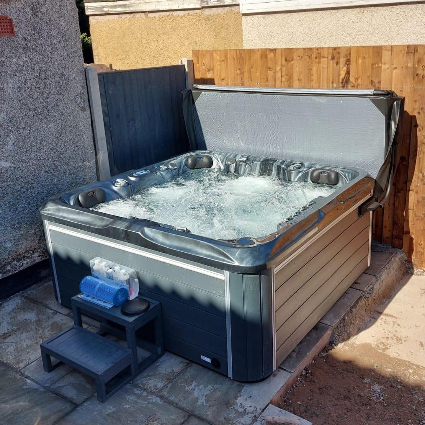 Hot tub with cover open, placed next to a fence. Water is bubbling, steps and towels are visible.
