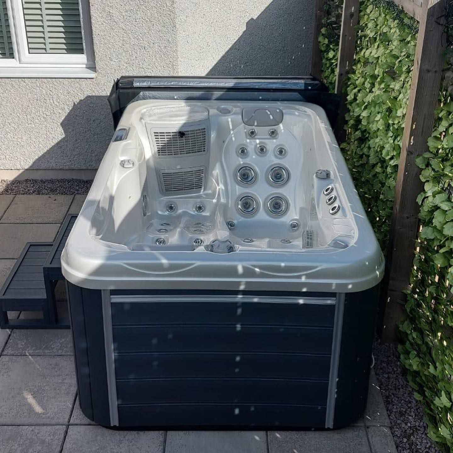 Hot tub on a patio, gray and white with dark blue skirting. Next to green foliage and a window.