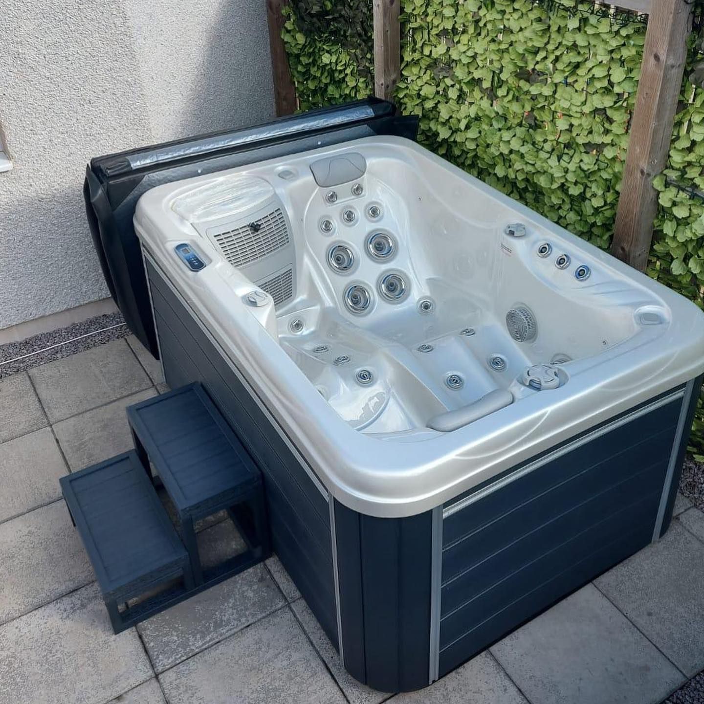 Hot tub on a stone patio with steps; cover open, surrounded by foliage, dark blue sides.
