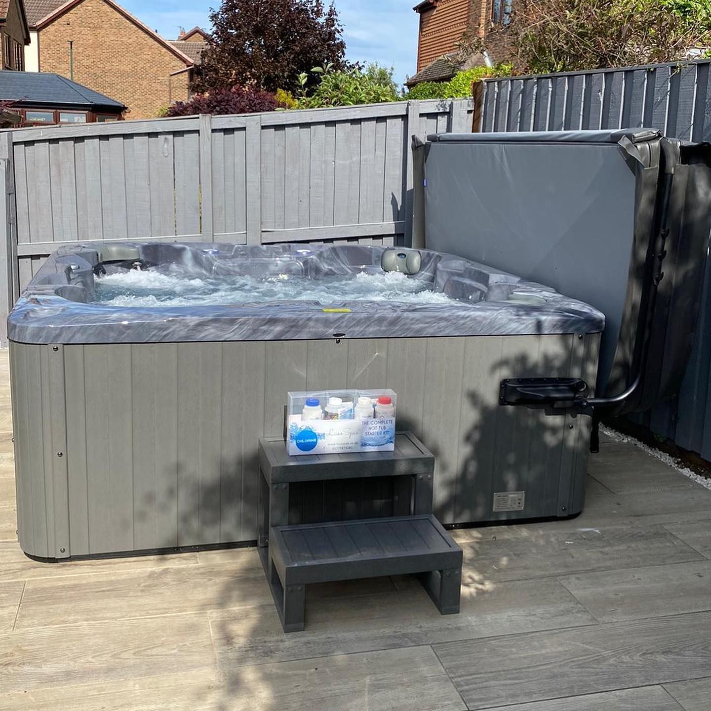 Gray hot tub with bubbling water in a backyard setting, open cover, small steps, chemical bottles.