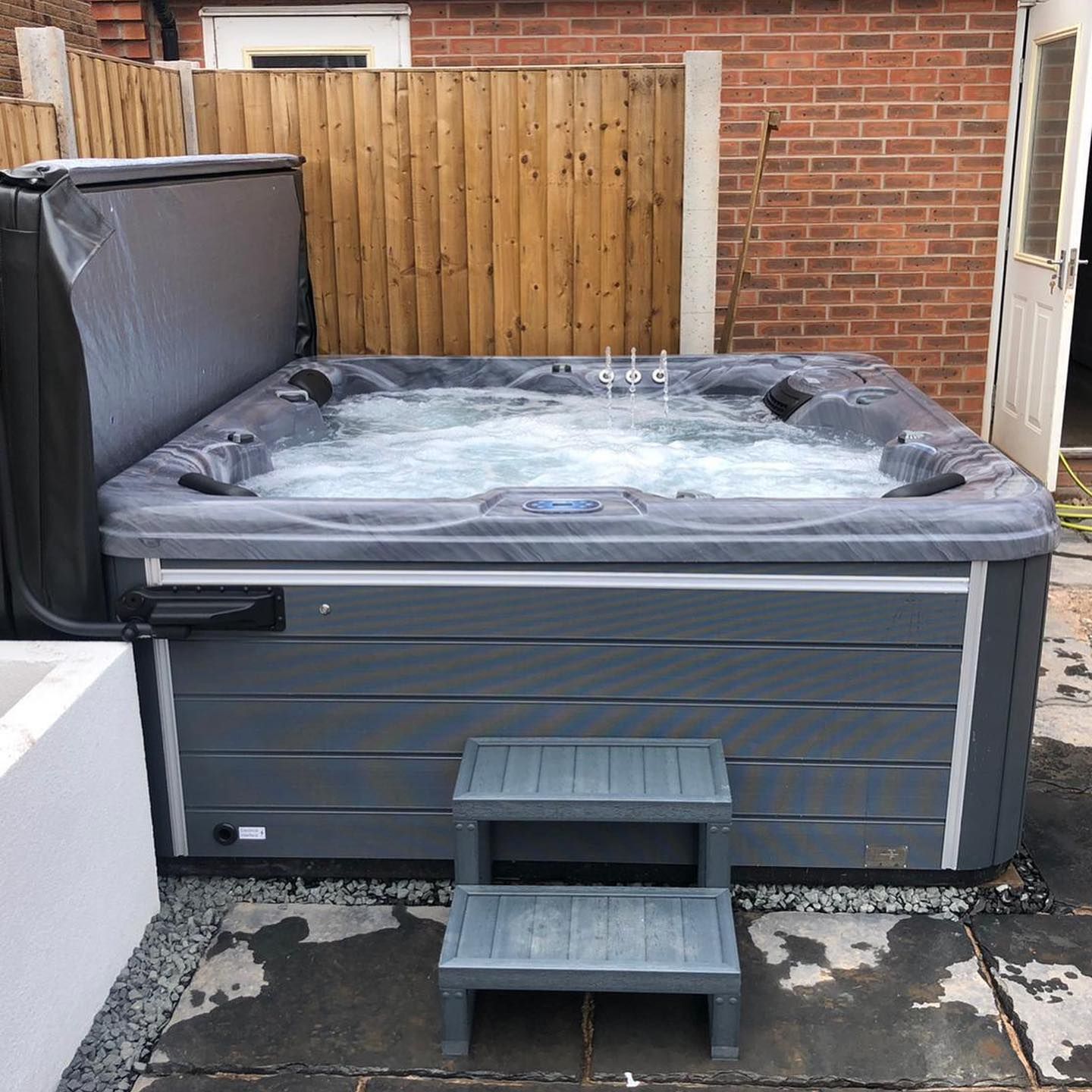 Gray hot tub with bubbling water, wooden steps, and a partially open cover, set outside.