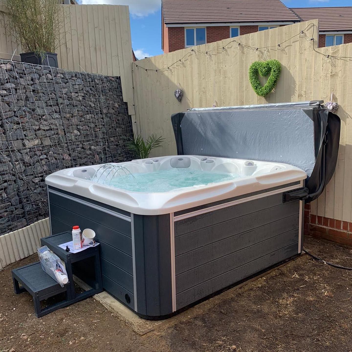 Hot tub in a backyard with stone wall and wooden fence. Gray hot tub cover open.