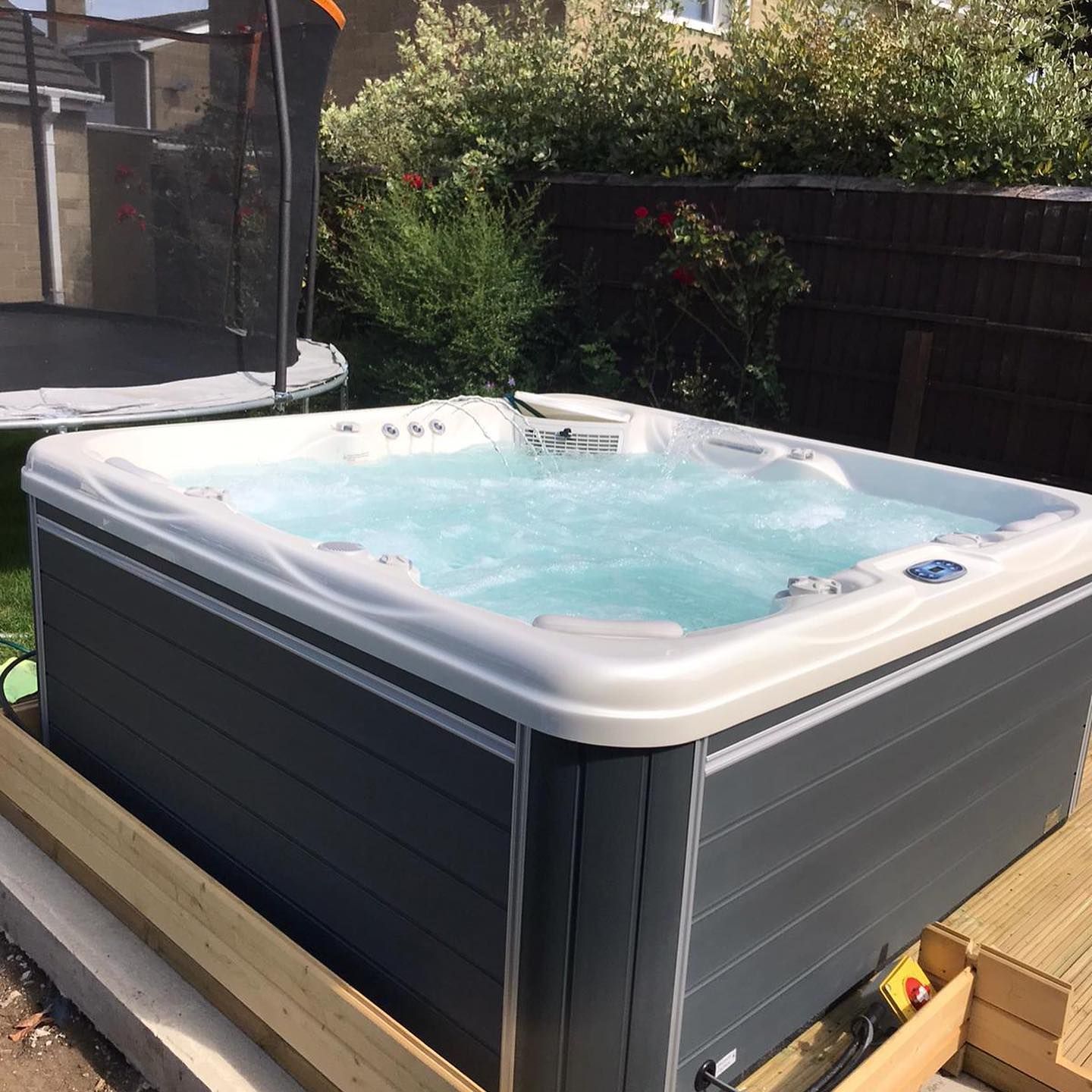 A hot tub in a backyard with a trampoline, surrounded by bushes and a wooden fence.