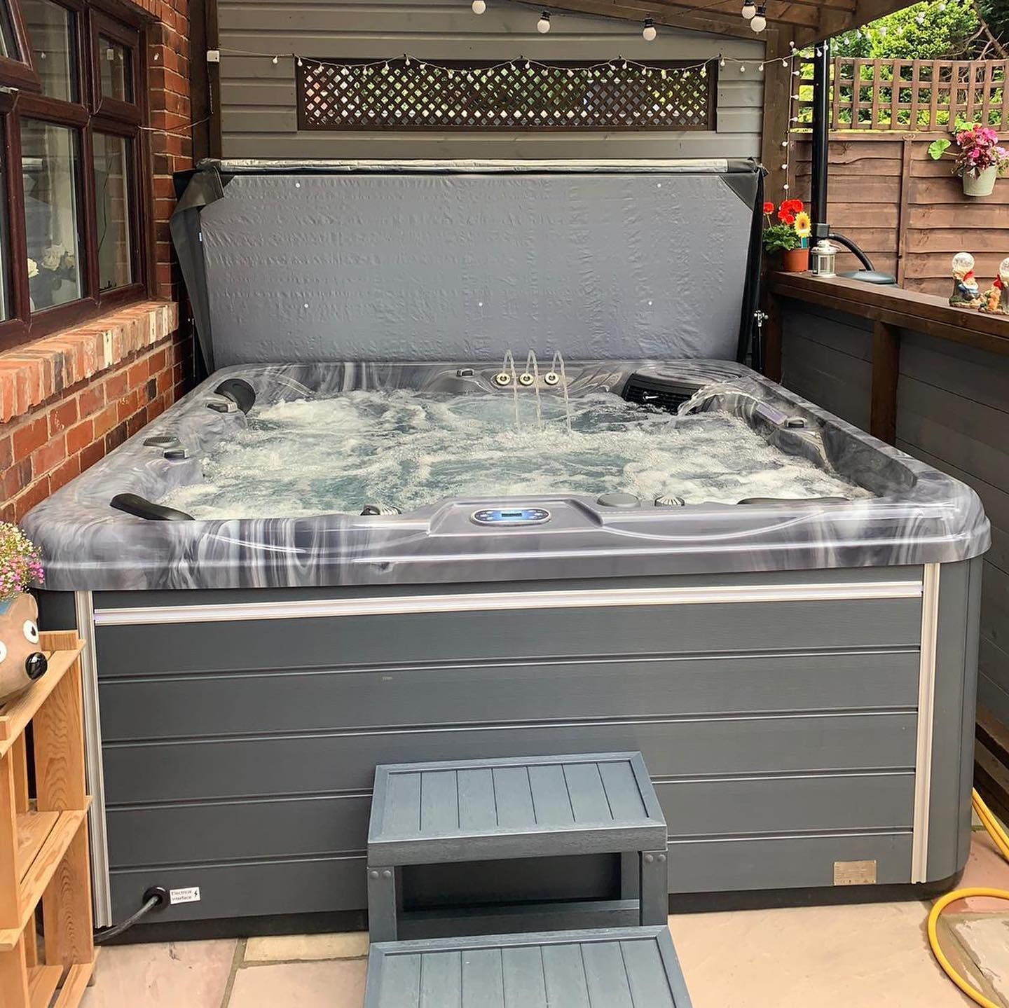 Hot tub with bubbling water, under a covered patio, next to a brick wall. Gray tub with steps.
