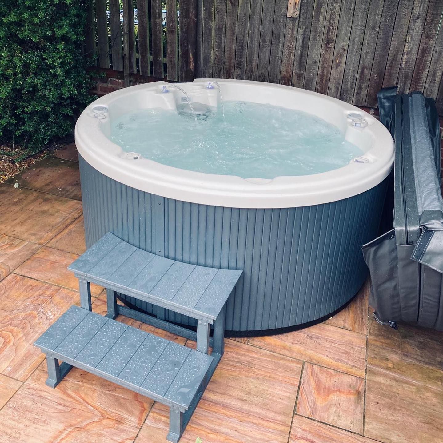 Round blue hot tub with steps, outdoors on a patio, partially covered with its grey cover.