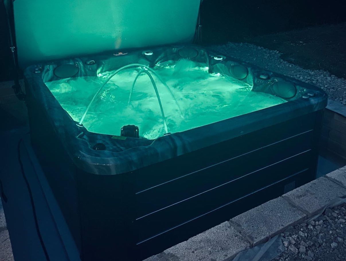 Lit-up green hot tub at night with water fountains. Black surround, open cover, set in brick surround.