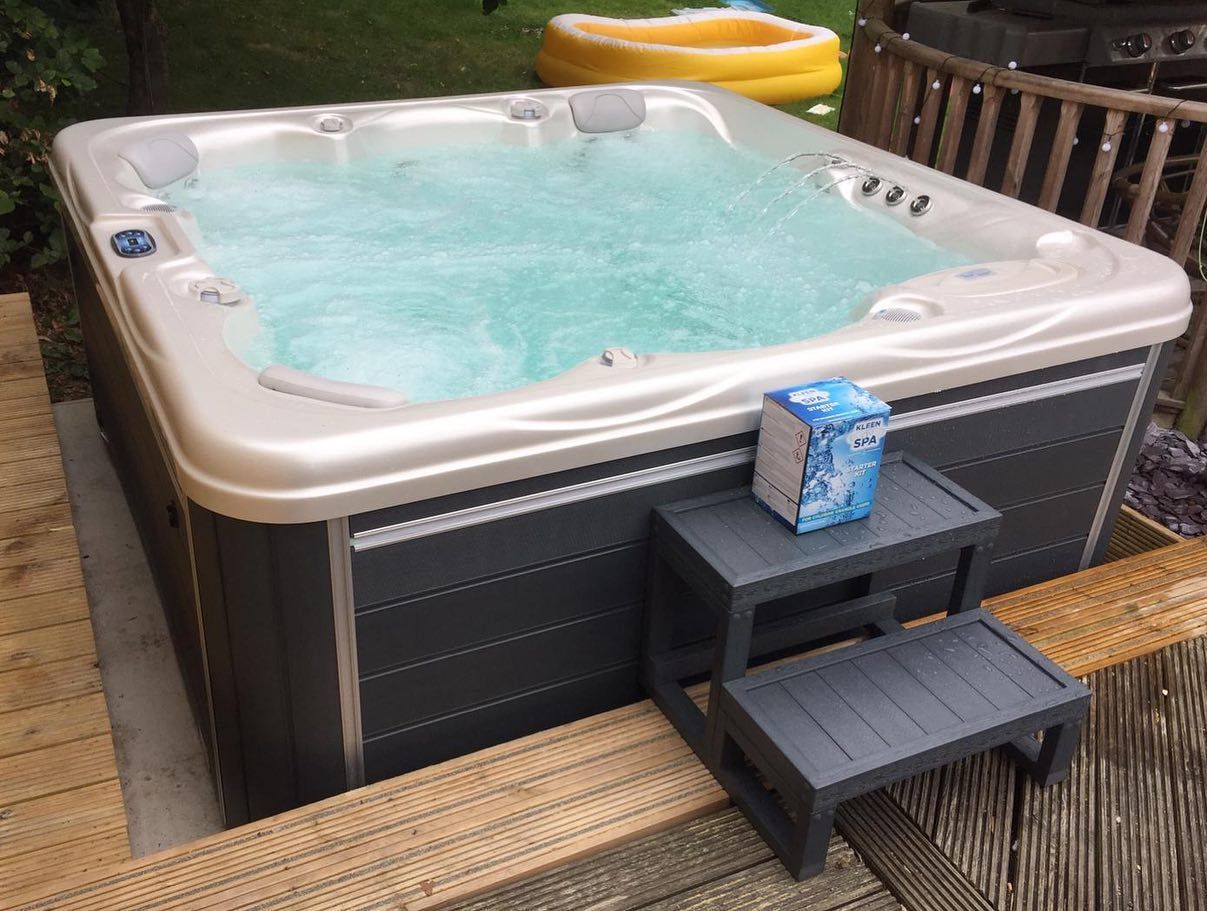 A hot tub filled with water on a wooden deck. A small step stool supports a box of chemicals.