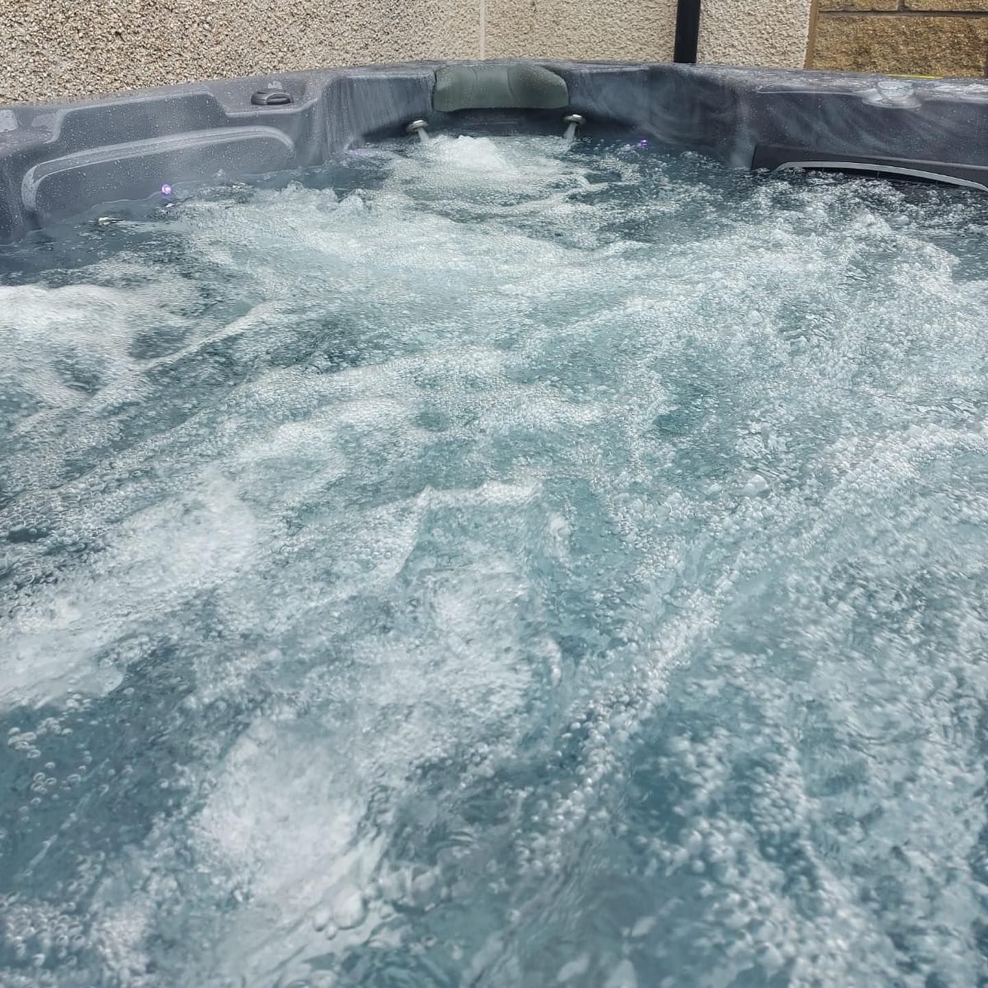 Bubbling, water-filled hot tub with jets creating white foamy water. Gray tub set against a beige wall.