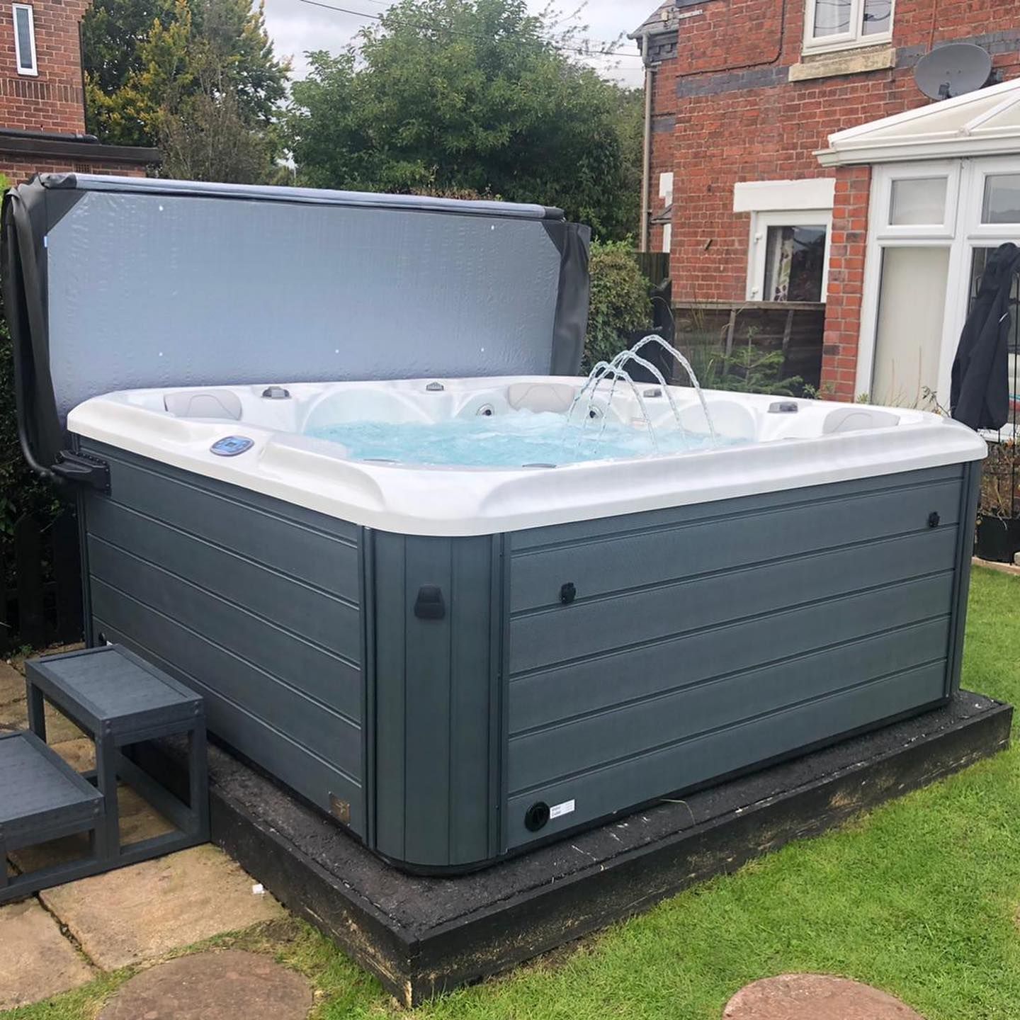 Dark gray hot tub with water jets, cover open, on a patio, surrounded by grass.