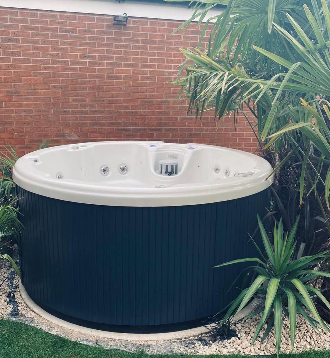 Round hot tub with dark blue exterior and white interior, set outside near plants and a brick wall.