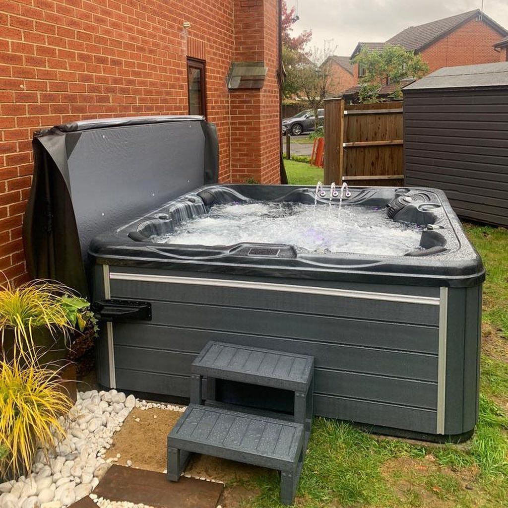 Dark gray hot tub with open cover, two-step entry, and water fountain jets, next to a brick wall.