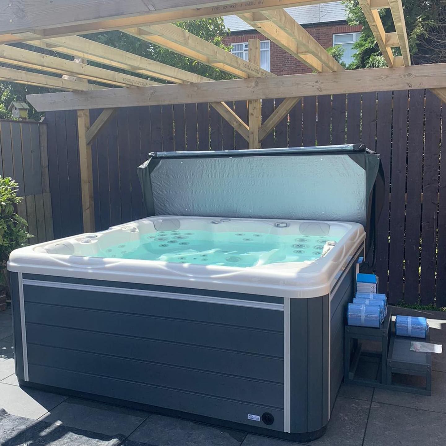 Hot tub with partially open cover, under a wooden pergola, in a backyard setting.