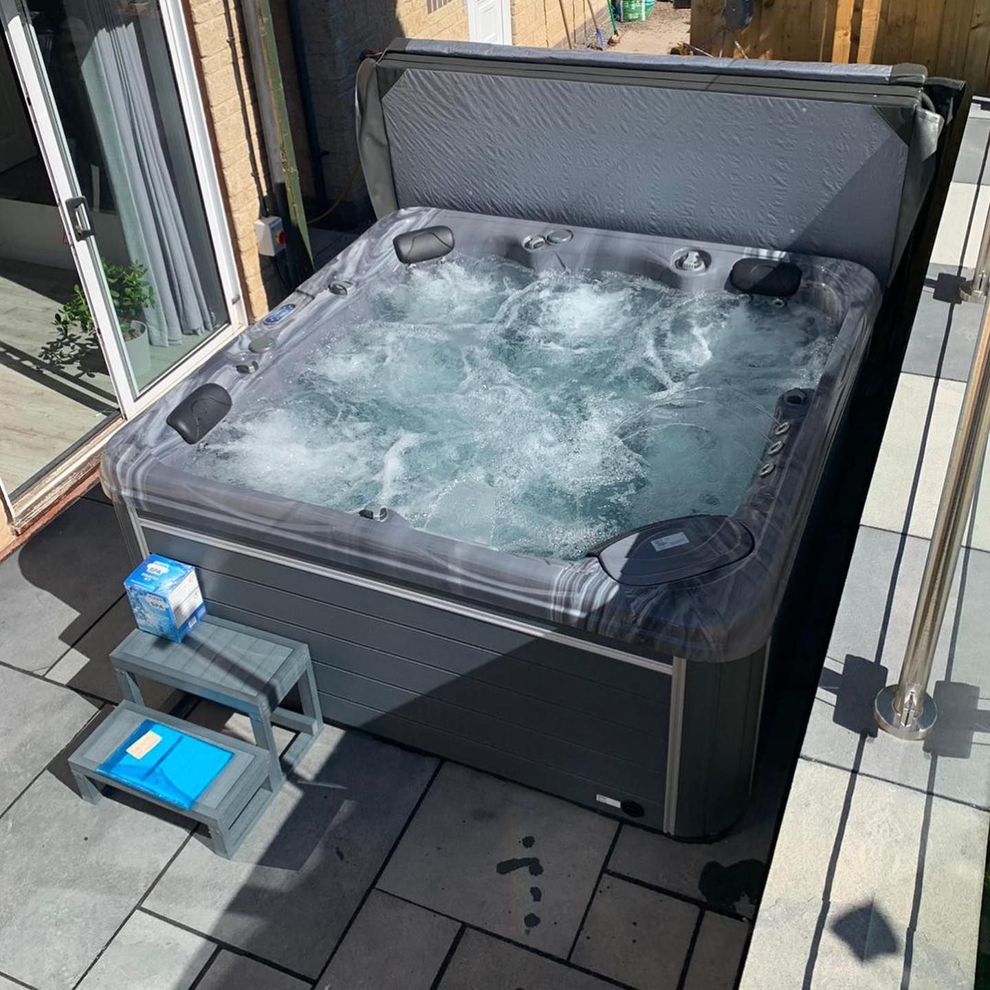 Hot tub on a patio with bubbling water, cover open, grey steps, and a sliding glass door nearby.