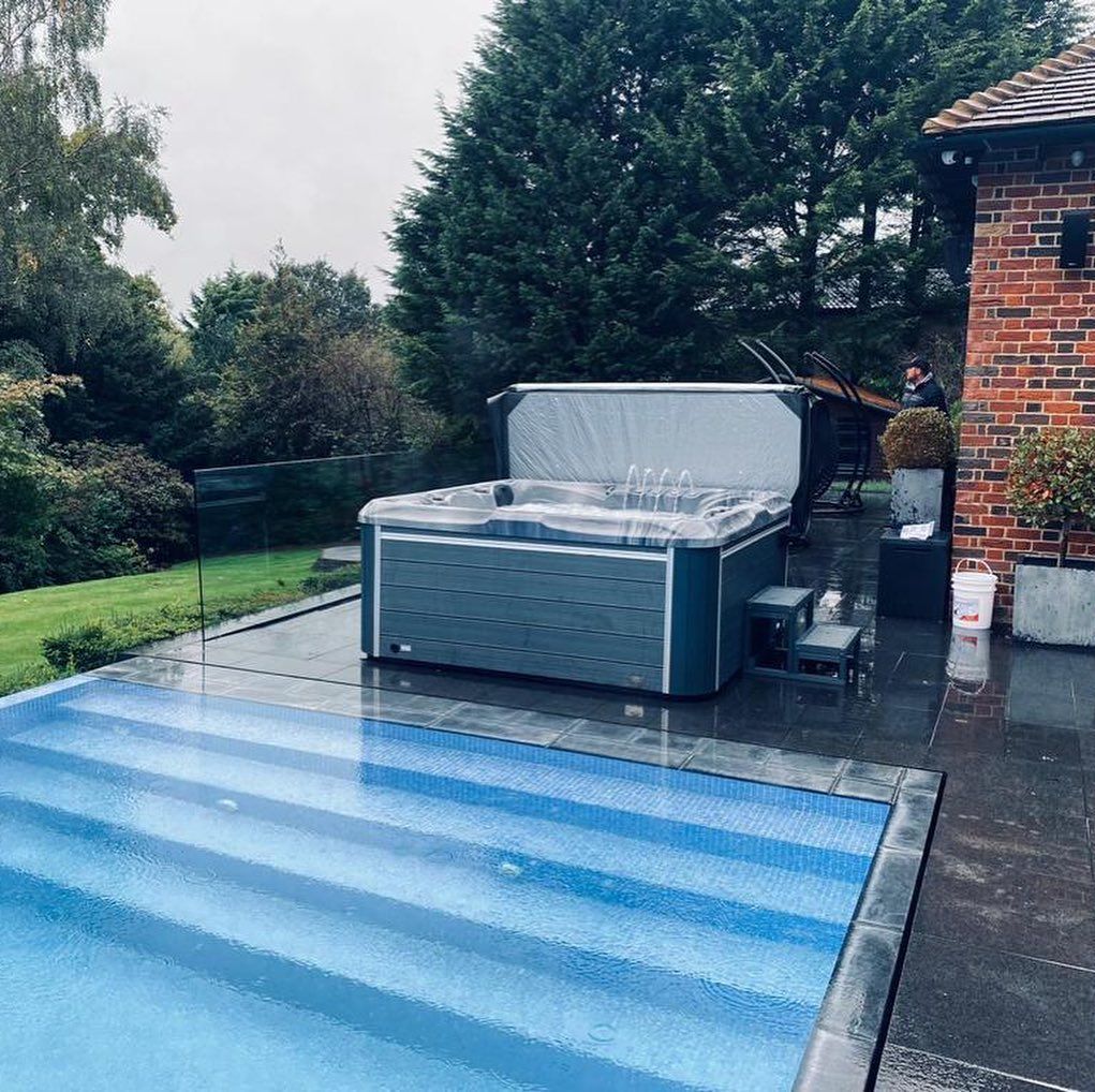 A hot tub and steps next to a swimming pool on a patio; trees and a brick building in the background.