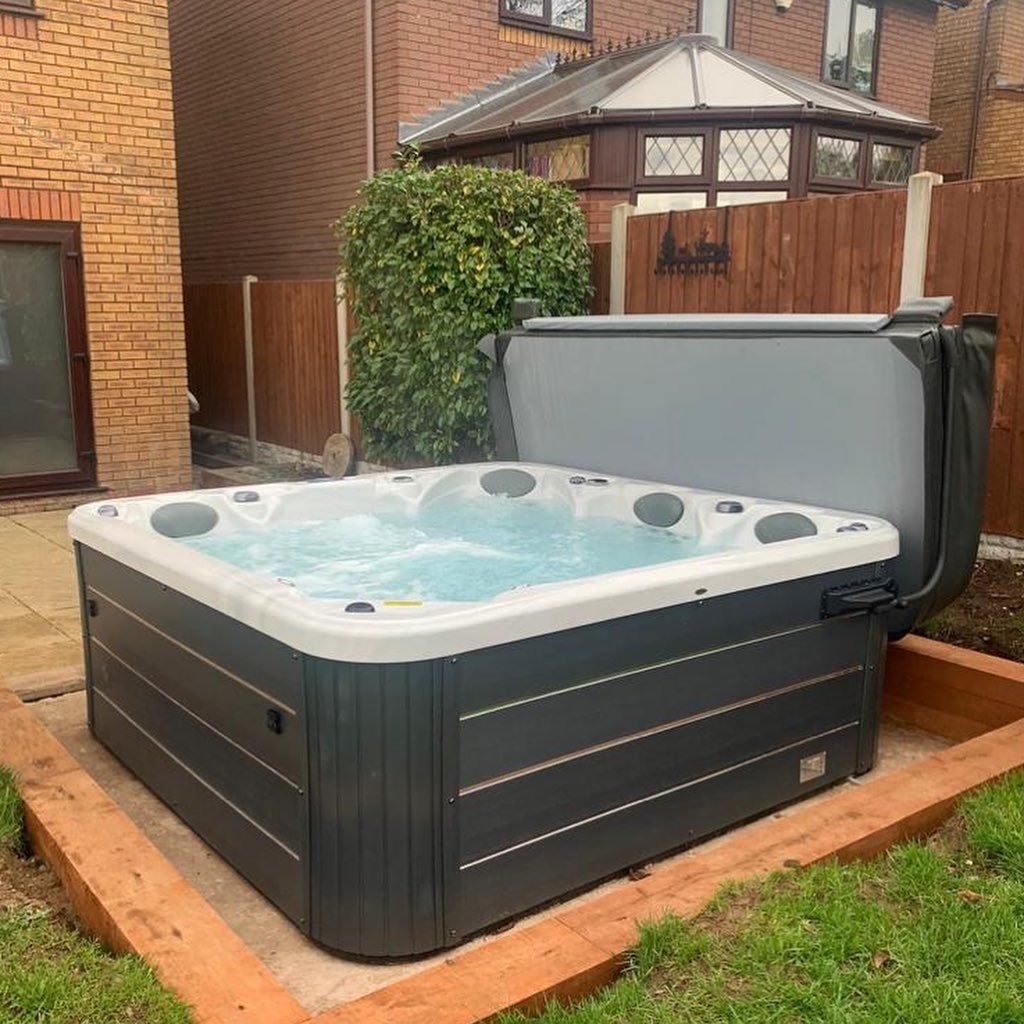 A square hot tub with the cover open sits in a backyard. The hot tub is filled with water.