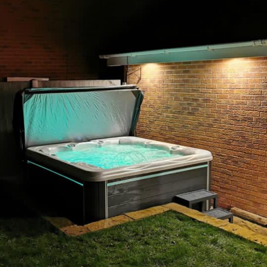 Lit hot tub outdoors at night, cover open, glowing aqua water and lights along the base, next to brick wall.