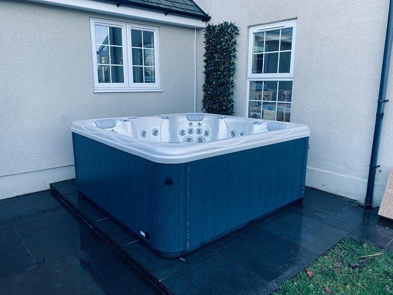 Blue hot tub on a dark stone patio, beside a building with white-framed windows.
