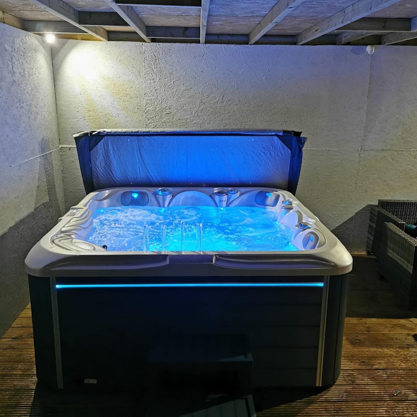 A lit hot tub with blue lights sits under a wooden deck, its cover partially open.