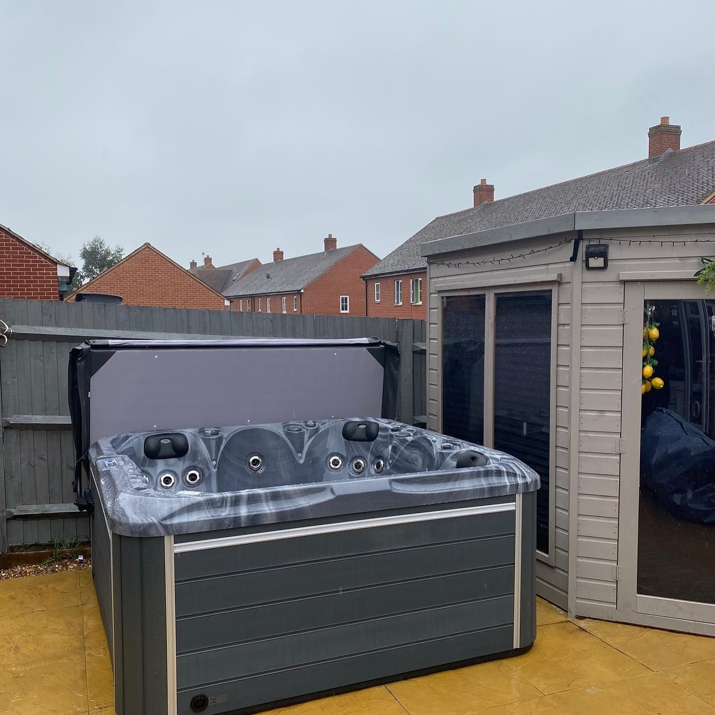 Hot tub with cover open next to a small shed in a backyard. Gray siding, brick homes in background.