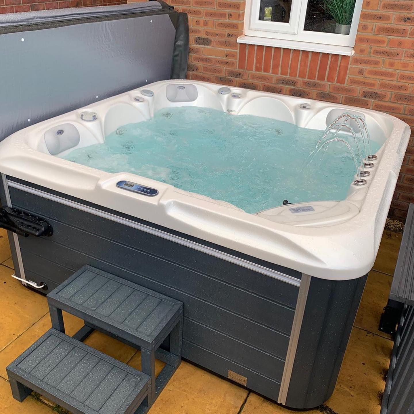 Hot tub filled with water; grey steps leading up to it.  Cover is partially open. Located outdoors.