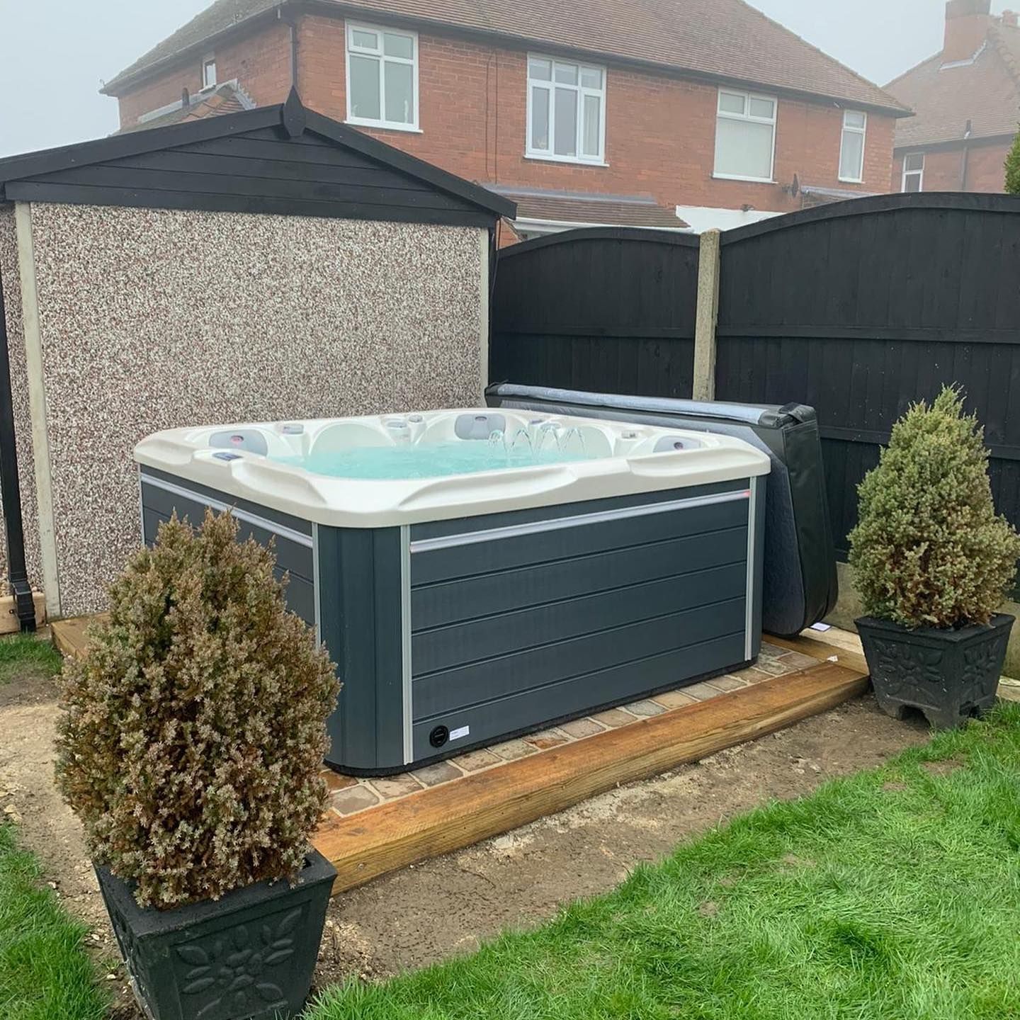 Hot tub surrounded by black fence and small trees.  Located next to shed in a backyard with grass.