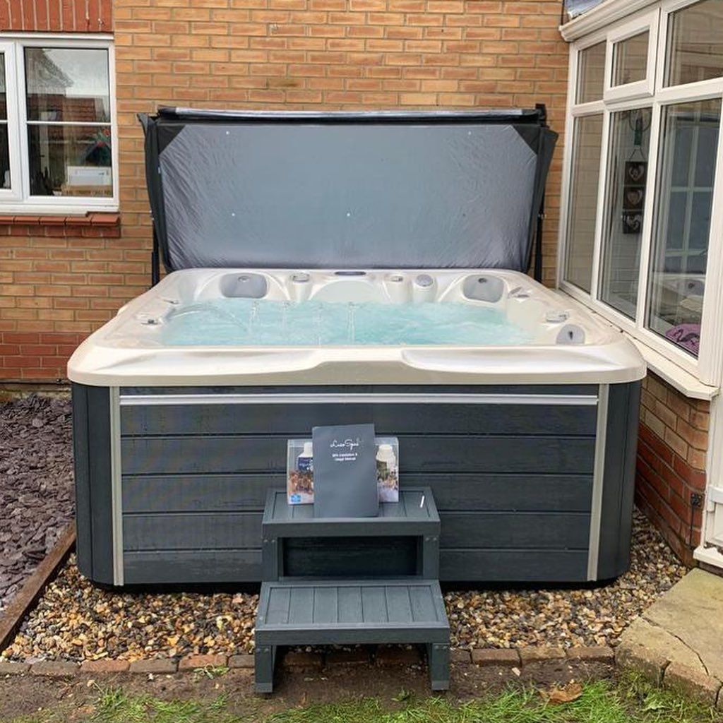 Hot tub with open cover, set on gravel, next to a house with window and step stool.