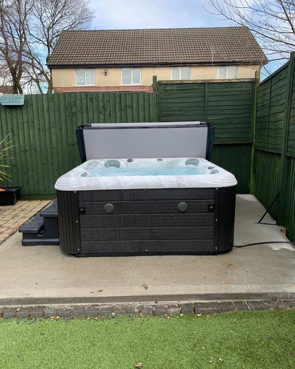 Hot tub with open cover on a concrete pad, surrounded by a green fence and grass.