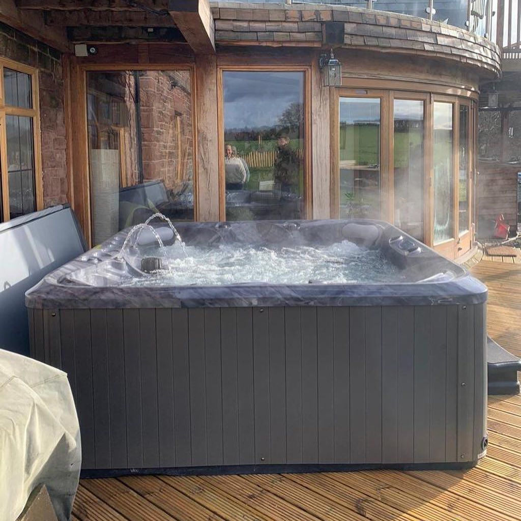 Hot tub on a wooden deck next to a building with glass doors and windows. Steam rises from the water.