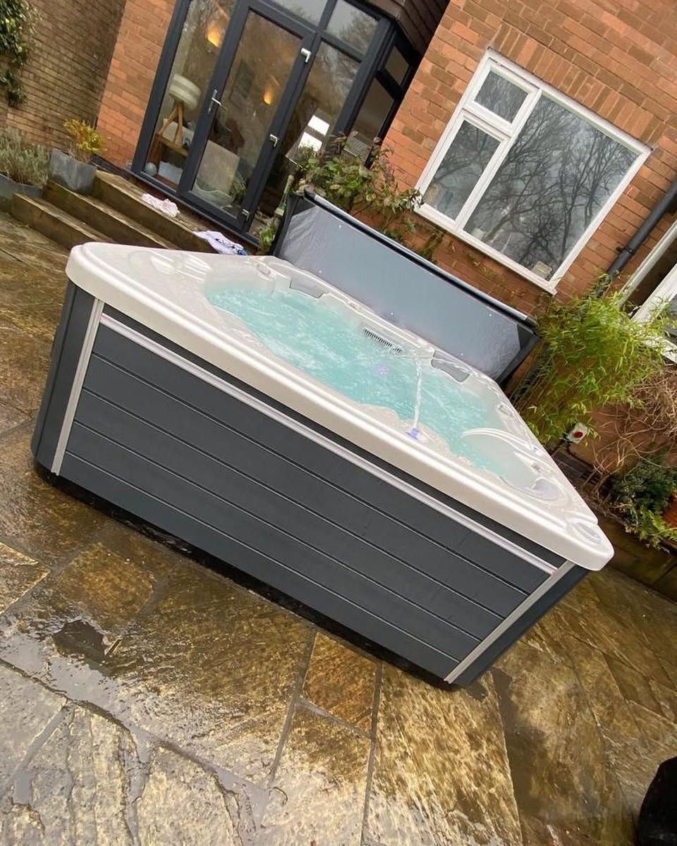 Hot tub with grey siding, set on a stone patio in front of a house.