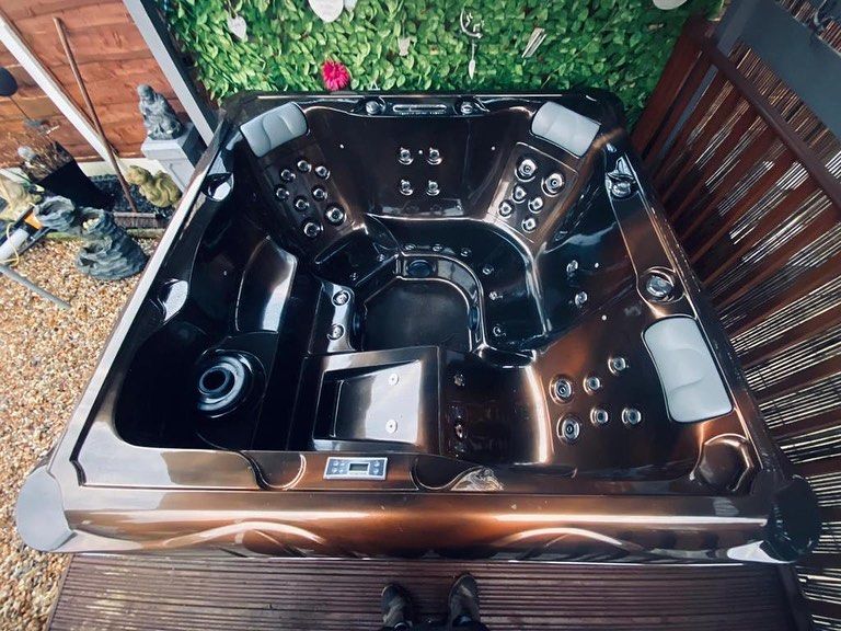 Brown hot tub, outdoor setting. Dark interior, multiple jets, surrounding wooden deck and greenery backdrop.