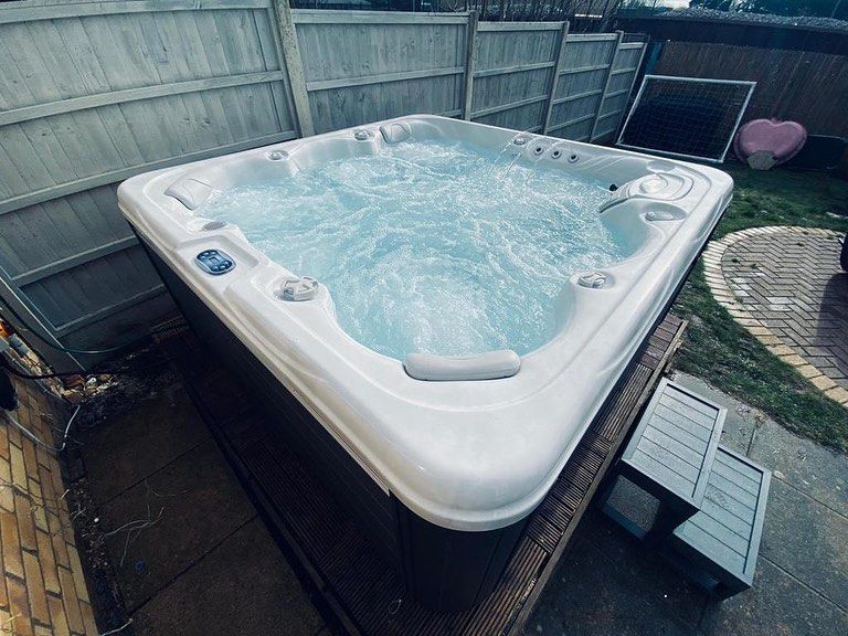 Square hot tub with bubbling water sits outdoors near a fence and brickwork.