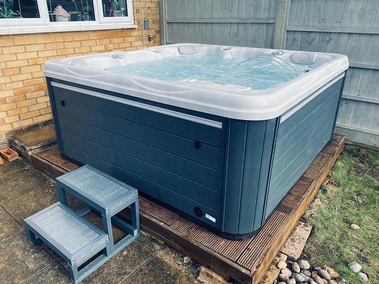 Hot tub on a wooden platform next to a step stool outdoors. Gray siding, white trim, brick wall.