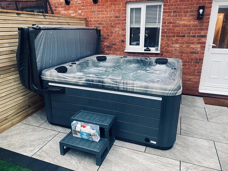 Hot tub with cover open, set on patio next to a brick wall. Gray steps lead to the tub.