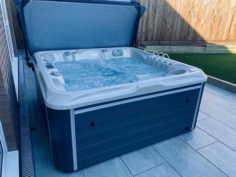 A hot tub with cover open, filled with water, sitting on patio tiles next to a fence.