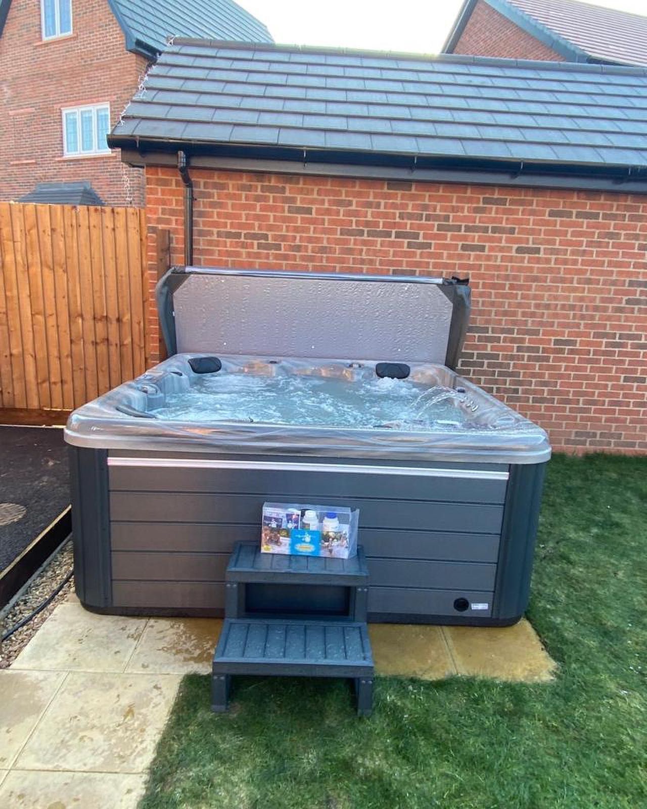 Hot tub in a backyard with brick wall, gray exterior, open lid, and a step stool.