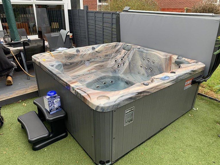 Hot tub on green turf, gray sides, marbled rim, open lid, steps to side, seated area behind.