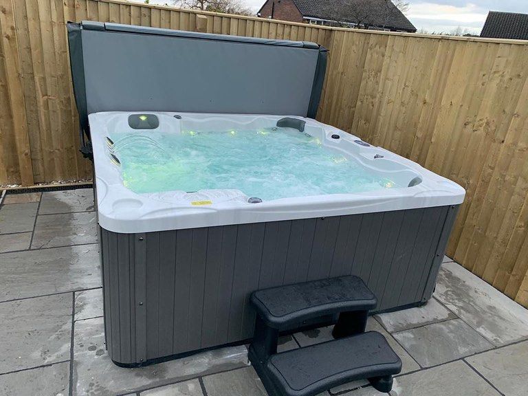 Hot tub with partially open gray cover, situated on gray patio tiles, with a gray step stool. Wooden fence backdrop.