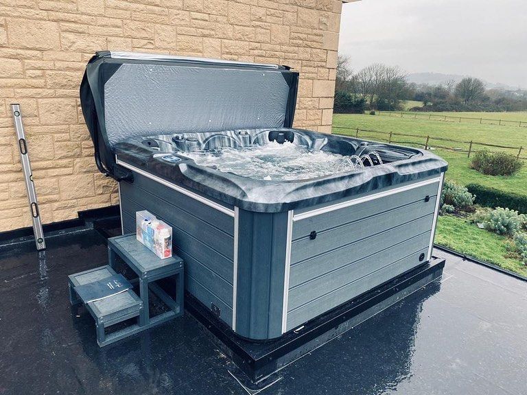 Hot tub with open cover on a dark platform, steps to the side, outside with rural scenery.