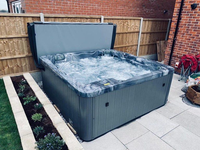 Gray hot tub with bubbling water sits in a backyard. Ajar cover. Landscaping surrounds it.