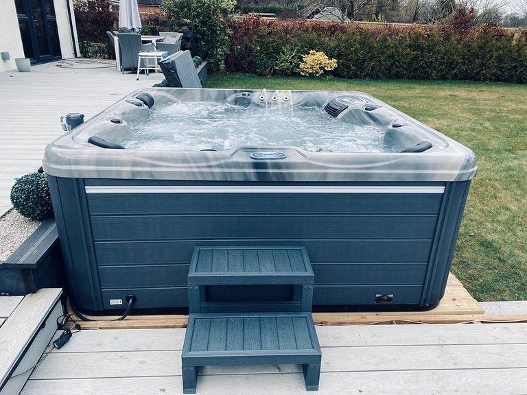 A square, gray hot tub with bubbling water sits on a wooden deck in a yard; a two-step stool is in front.